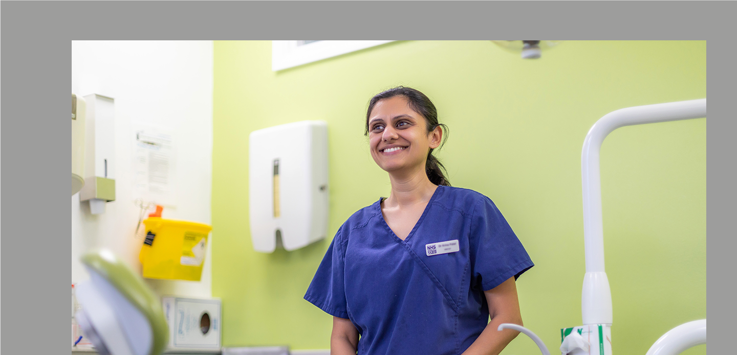 A dentist wearing navy blue scrubs smiles standing in front of a lime green wall in a surgery